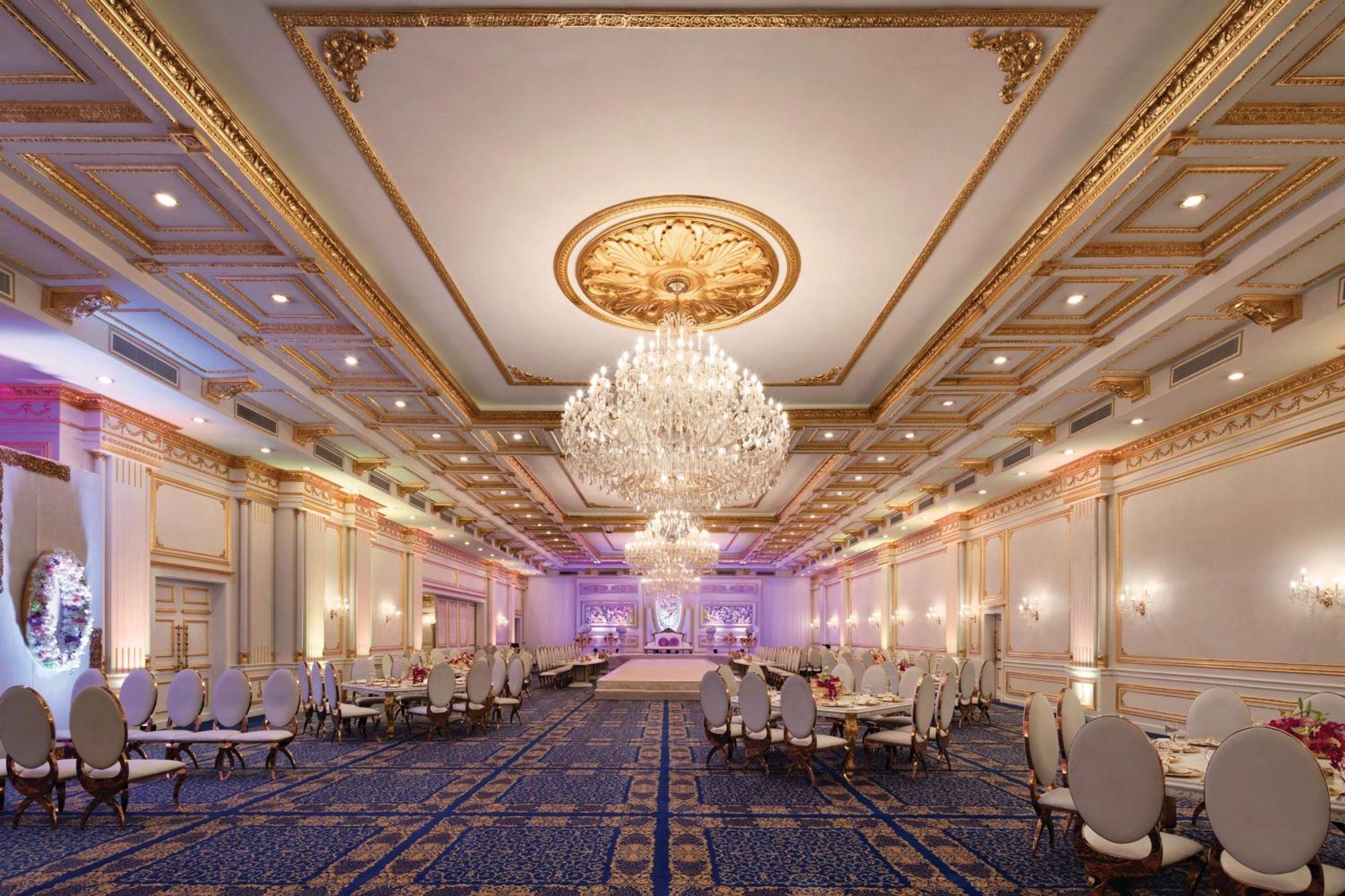 Grand Regency Doha, Trademark Collection By Wyndham Dış mekan fotoğraf The photo shows an elegant banquet hall with luxurious decor. The room features high ceilings adorned with intricate gold details and a large chandelier that hangs in the center, providing soft illumination. There are several round tables with plush 