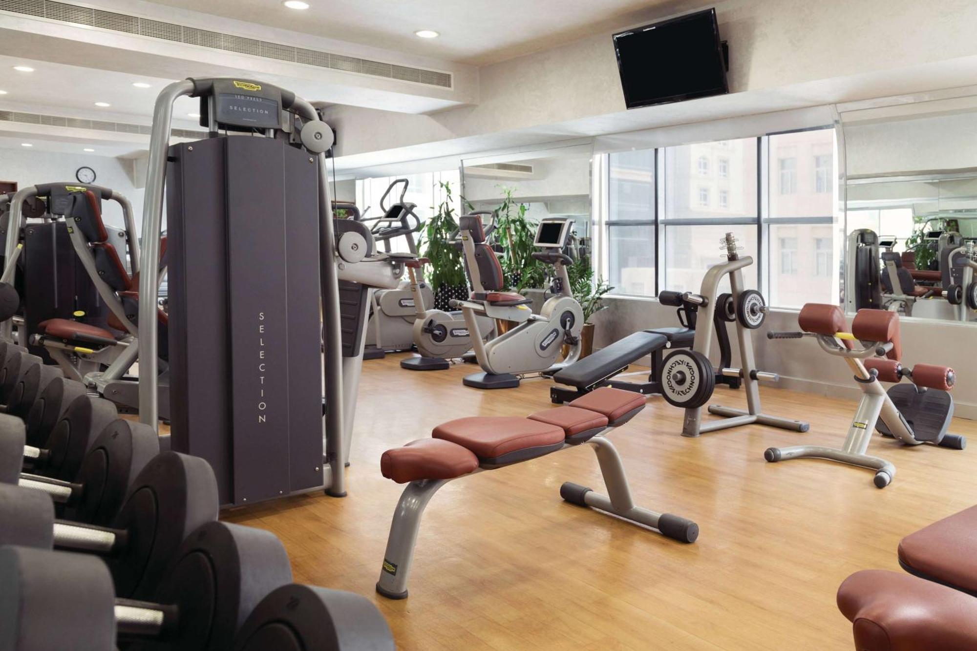 Grand Regency Doha, Trademark Collection By Wyndham Dış mekan fotoğraf The photo shows a fitness gym with various exercise equipment. There are weight machines, benches, and cardio machines such as stationary bikes and treadmills. The space has large windows allowing natural light to enter, and there are mirrors on the 