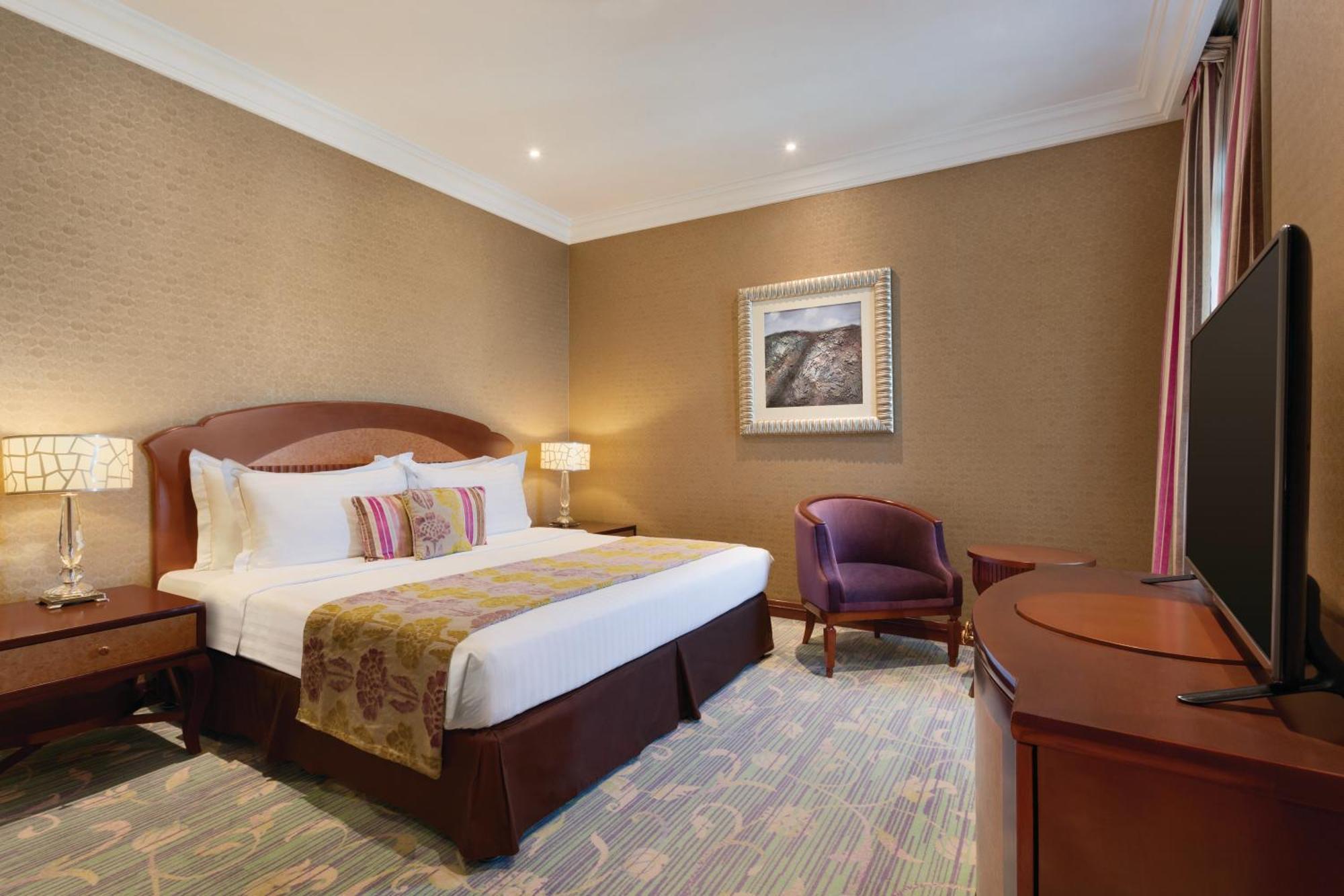 Grand Regency Doha, Trademark Collection By Wyndham Dış mekan fotoğraf The image shows a hotel room that features a cozy and elegant design. There's a large bed with white sheets and colorful decorative pillows. Next to the bed, there are nightstands with lamps. A comfortable armchair is positioned in the corner, and a 
