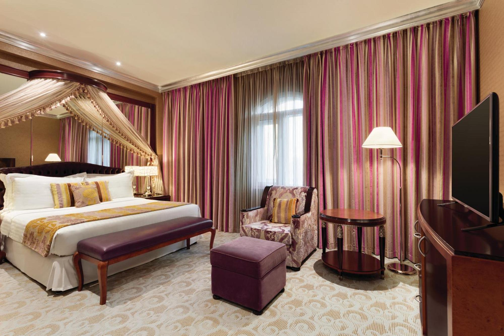 Grand Regency Doha, Trademark Collection By Wyndham Dış mekan fotoğraf The photo shows a stylish hotel room. It features a large bed with decorative pillows and a draped canopy above it. The room has elegant striped curtains in shades of pink and purple. There is a small seating area with a plush chair and ottoman, alon