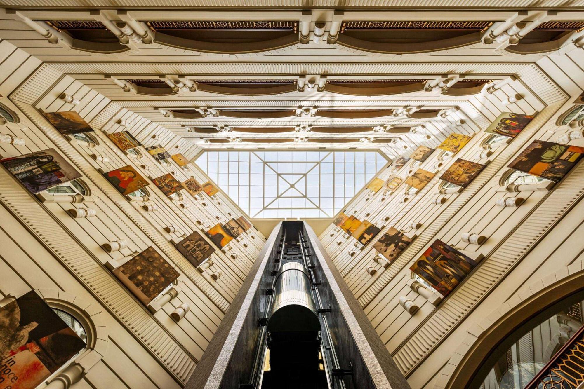 Grand Regency Doha, Trademark Collection By Wyndham Dış mekan fotoğraf The photo shows an interior view of a building, captured from a low angle looking upwards. The architectural design features a tall, open space with walls adorned with various artworks. In the center, there is a glass ceiling that allows natural ligh
