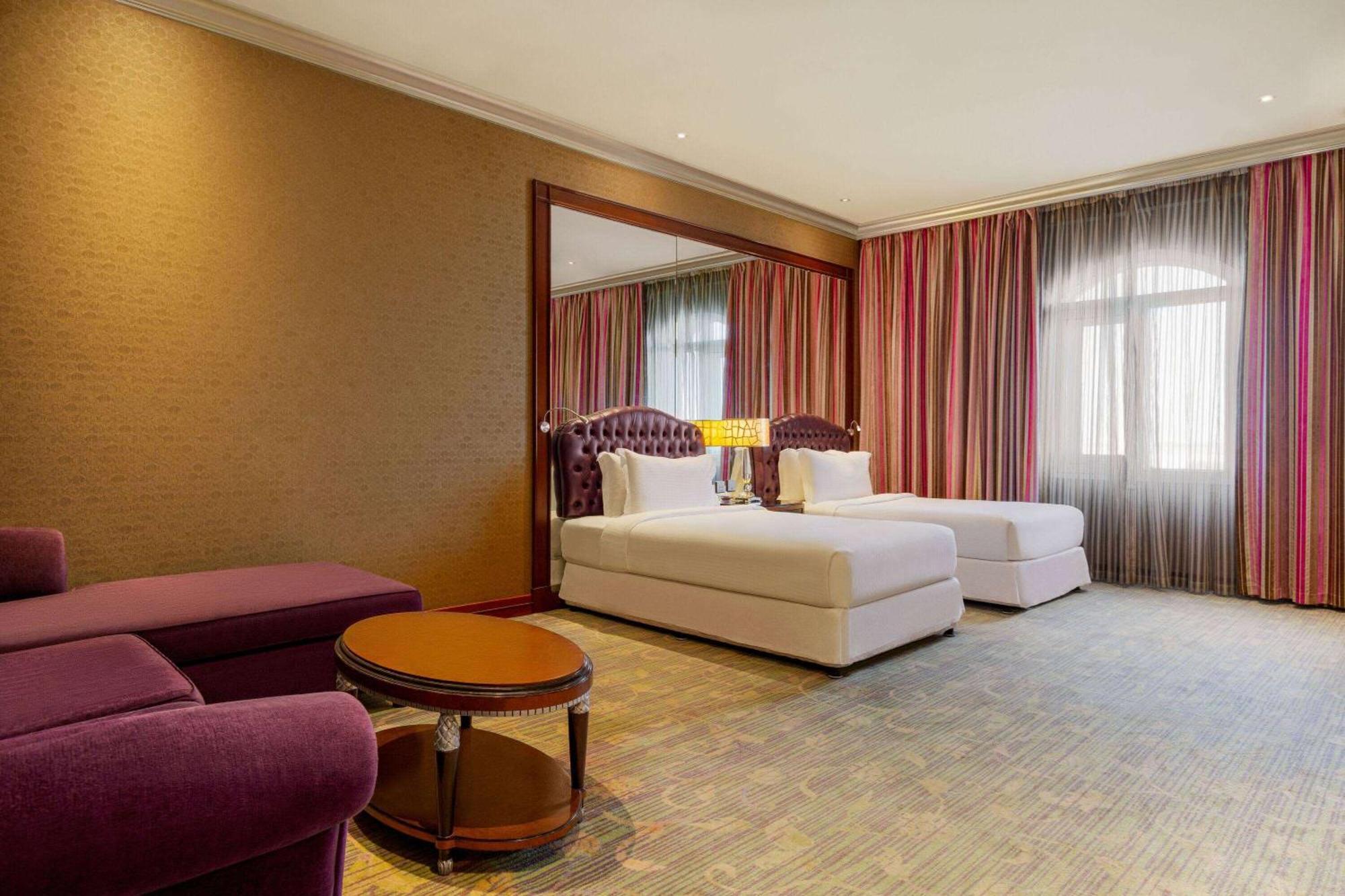 Grand Regency Doha, Trademark Collection By Wyndham Dış mekan fotoğraf The photo shows a hotel room featuring two beds with white bedding, a large mirror on one wall, and a window dressed with sheer and curtain panels. There is a small round table in the center of the room and a plush purple armchair nearby. The overall