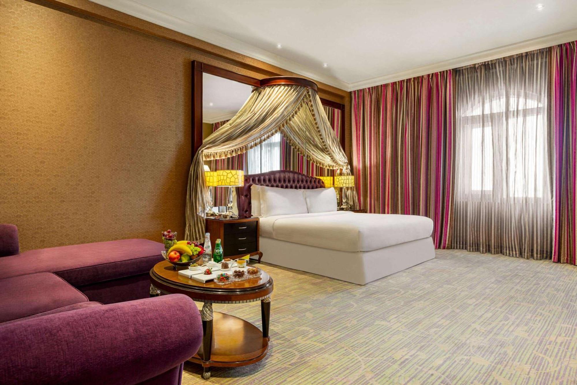 Grand Regency Doha, Trademark Collection By Wyndham Dış mekan fotoğraf The photo shows a luxurious hotel room featuring a king-sized bed with a decorative canopy. The room is elegantly designed with a mix of rich colors and textures. To the left, there's a plush purple seating area with a small round table that displays
