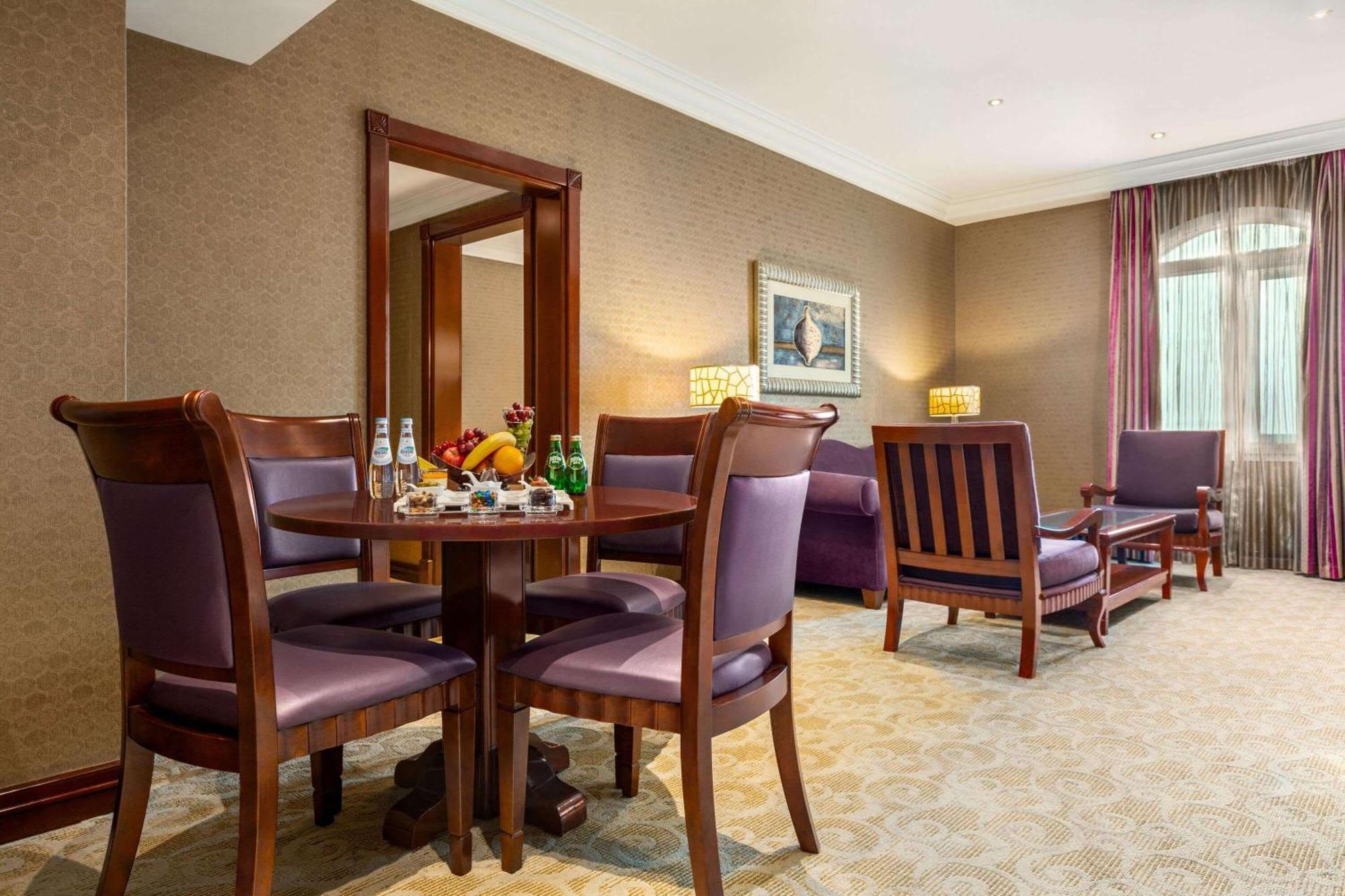 Grand Regency Doha, Trademark Collection By Wyndham Dış mekan fotoğraf The photo depicts a well-furnished hotel room or lounge area. In the foreground, there is a round table surrounded by four purple upholstered chairs, with a selection of fruits and beverages on the table. To the right, there are two wooden armchairs,